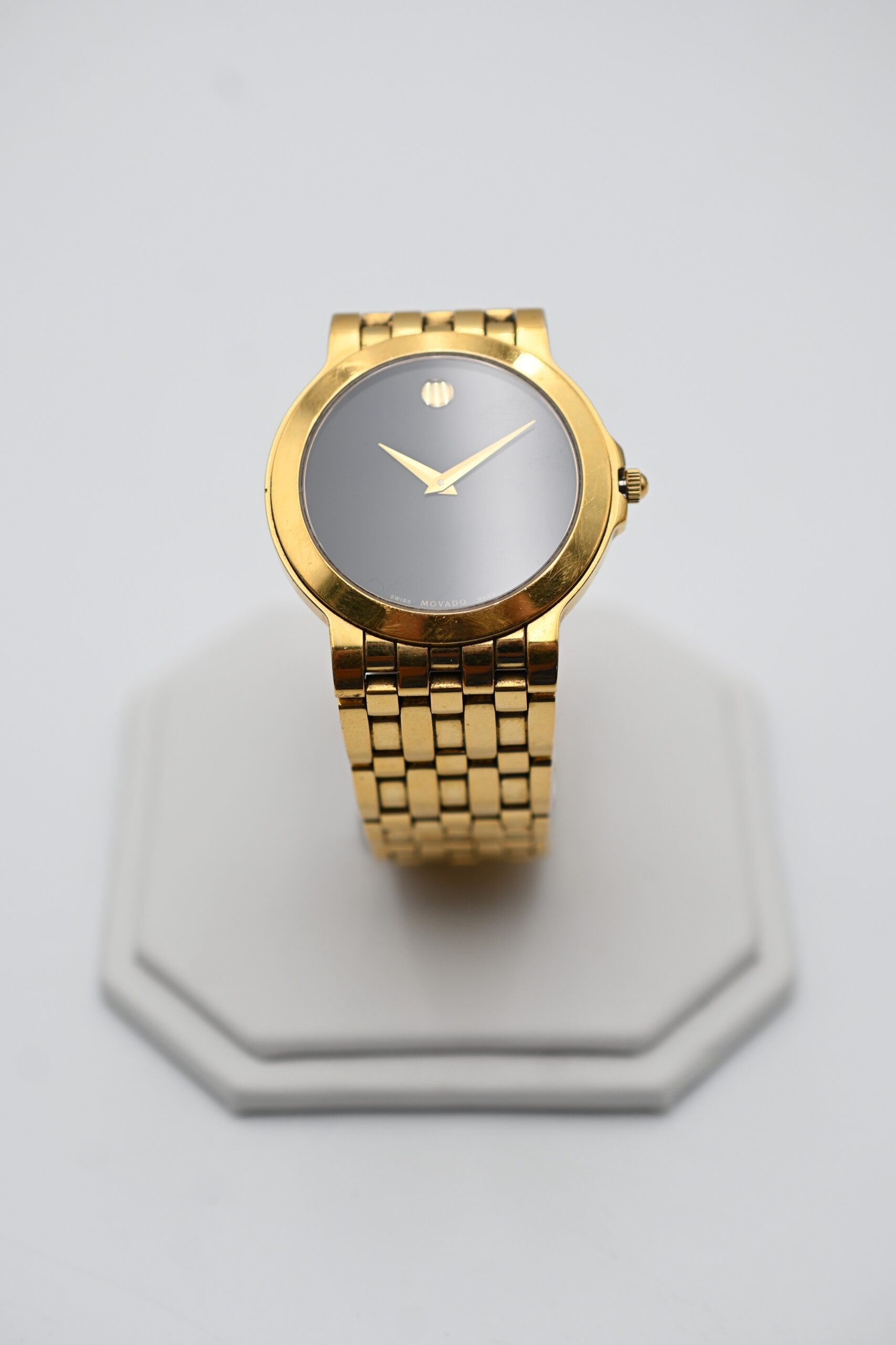 Gold-tone Movado wristwatch displayed upright on a jewelry stand, showing the Museum dot dial